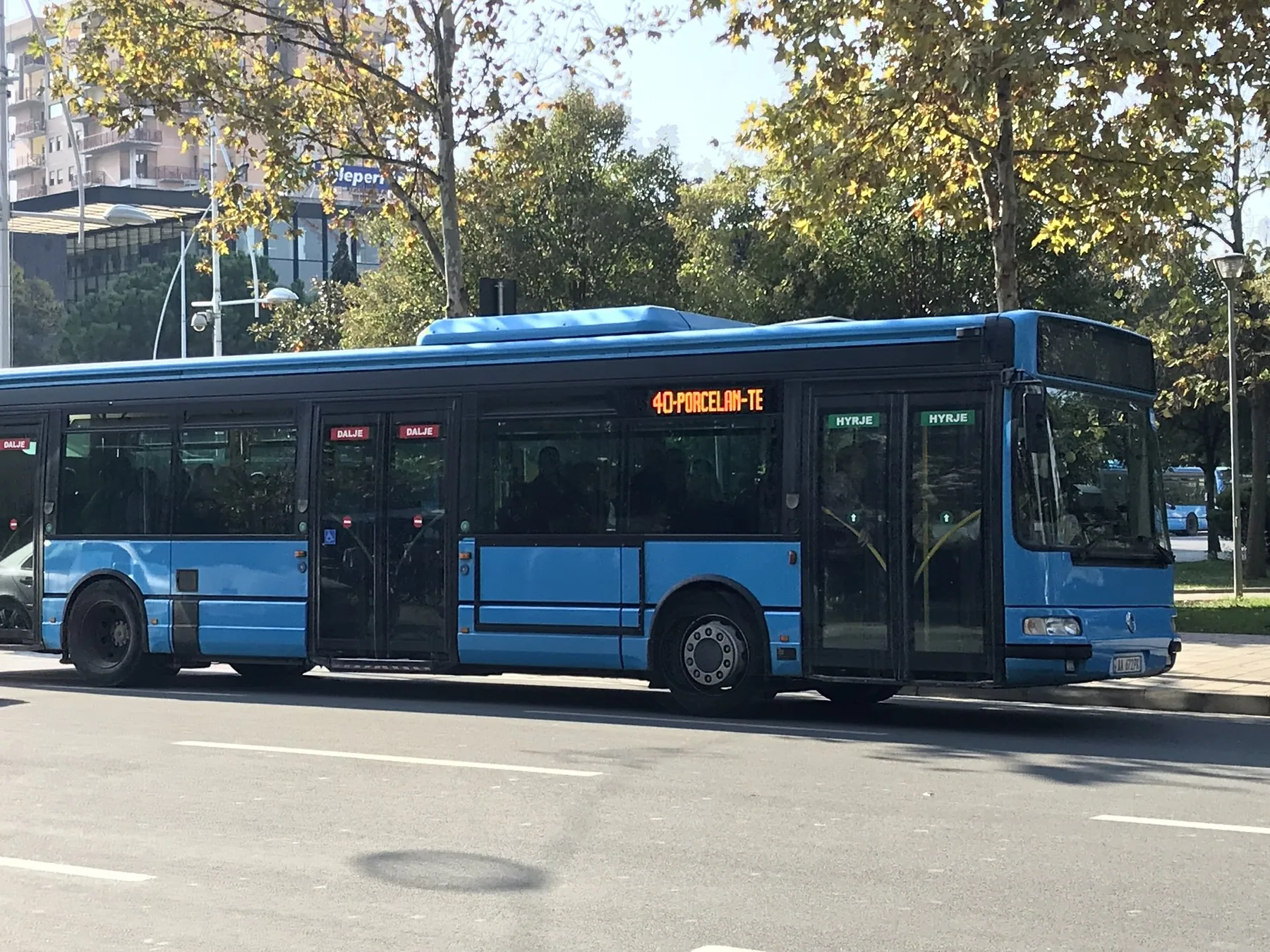 Blå stadsbuss på en gata i Tirana med lövträd och bostadshus i bakgrunden