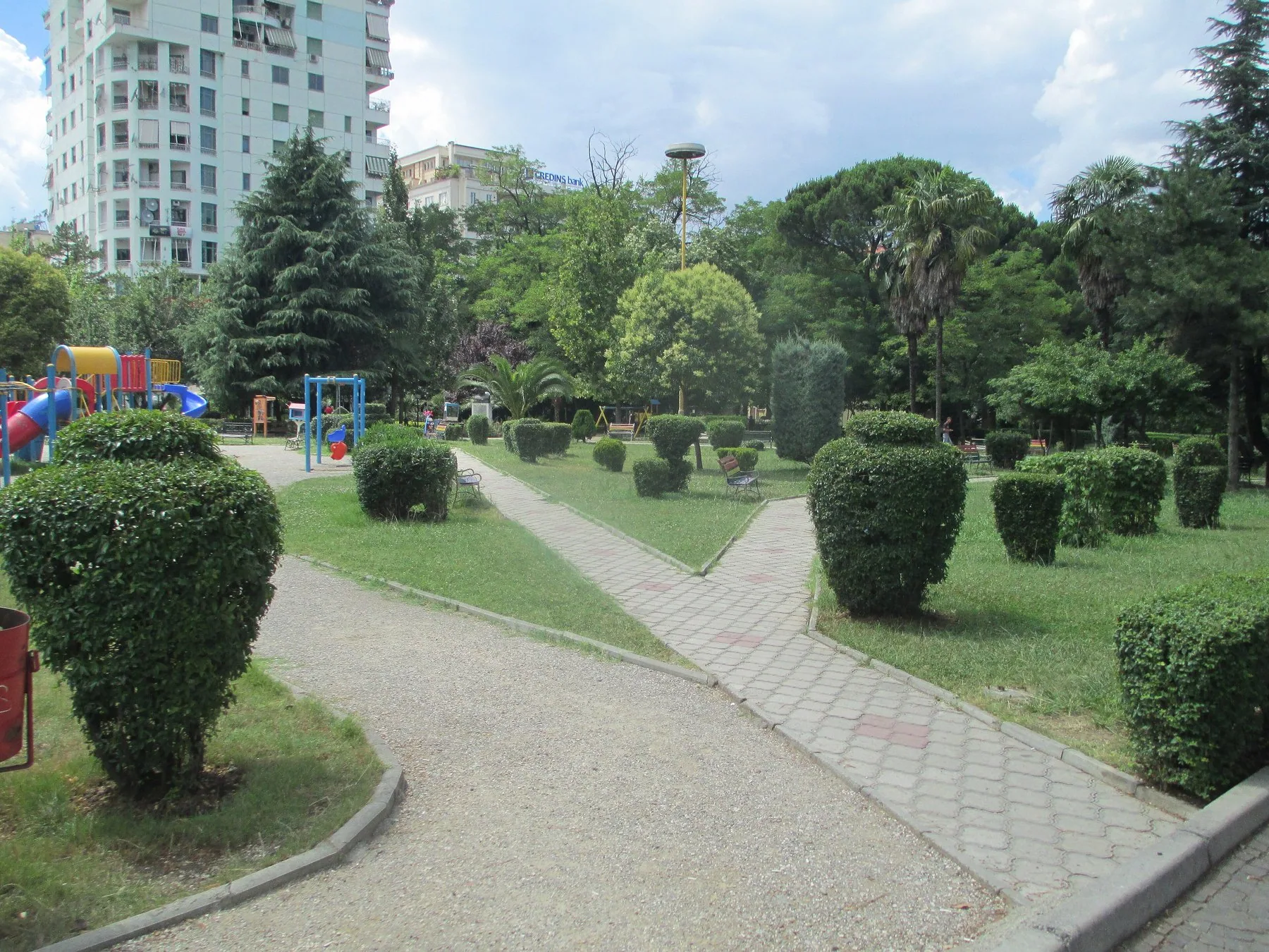 Lekplats med färgglada klätterställningar bland klippta buskar och gångar i en park i Tirana